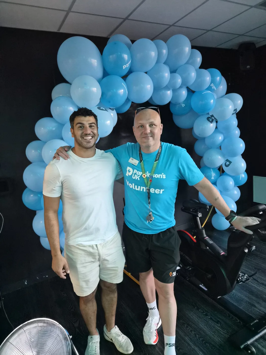 Two men in front of an arch of light blue Parkinson's UK balloons beside an exercise bike, one in a Parkinson's UK volunteer T-shirt