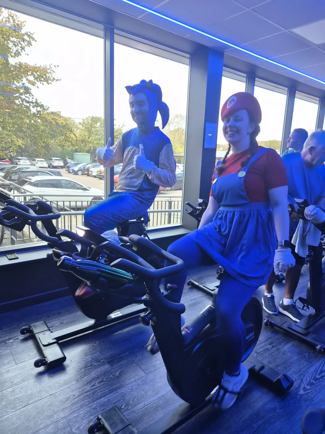 Two riders on exercise bikes, one dressed as Sonic the Hedgehog giving a thumbs up and one dressed as Mario